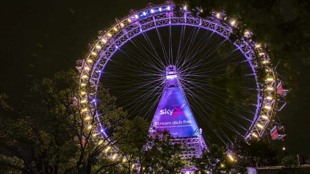 Riesenrad bei Nacht mit bunten Lichtern, im Hintergrund der Himmel, beworben für Sky X.